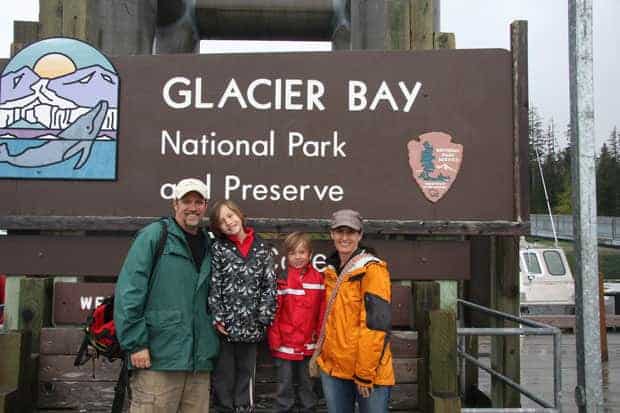 A family of four stands in front of the brown wooden sign for Glacier Bay Alaska National Park