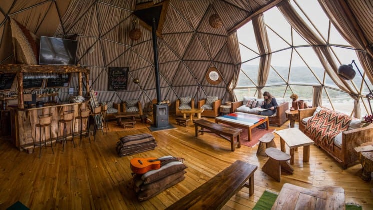A woman reads by the window in the Community Dome with couches and chairs, tables and a five-stool bar at EcoCamp in Patagonia, Chile