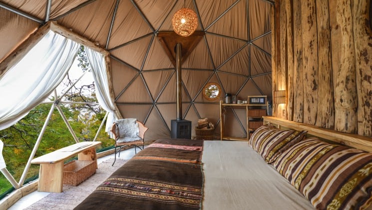 Interior of dome with large bed, small stove and open curtains showing surrounding forest at EcoCamp in Patagonia, Chile