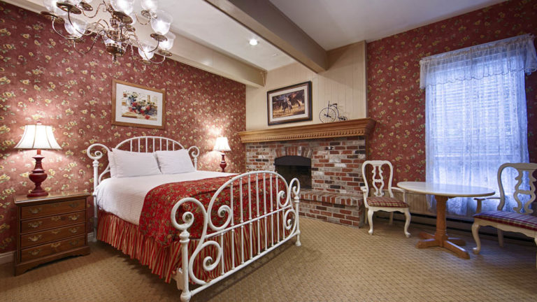 A room with a metal Victorian-style bed frame, red linens, a brick fireplace, and a chair at the Best Western Grandma's Feather Bed Hotel in Juneau, Alaska