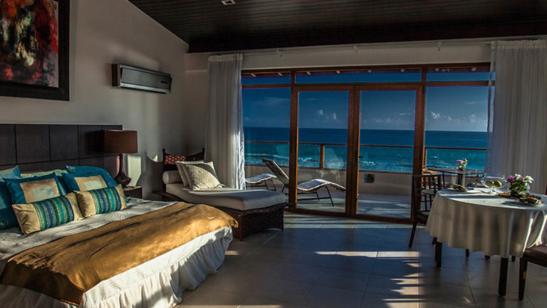 The light fades on the master suite at Iguana Crossing Boutique Hotel at dusk, with a king-sized bed, a sliding glass door, and a balcony overlooking the ocean in the Galapagos Islands