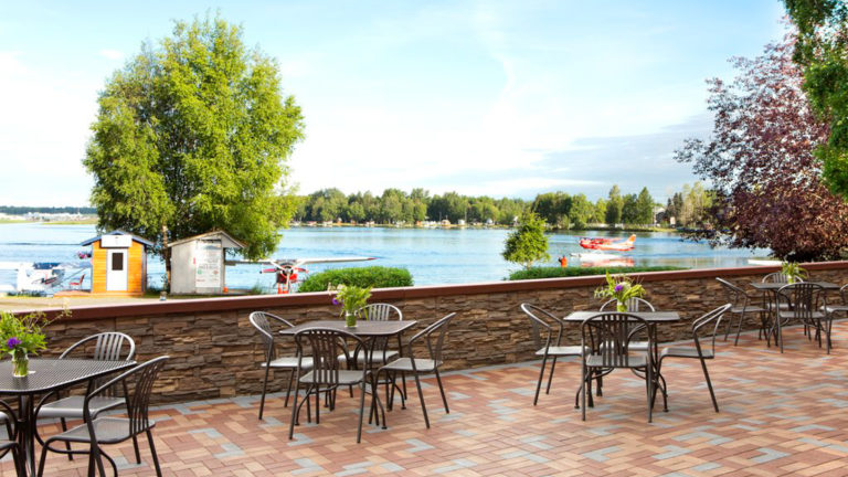 Tables are set on the patio in front of the Lakefront Anchorage, a hotel in Alaska, with a view of the water.