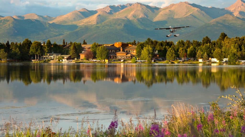 At dusk, a plane flies above the Lakefront Anchorage Lodge, with mountains in the background and water reflecting trees in the foreground.