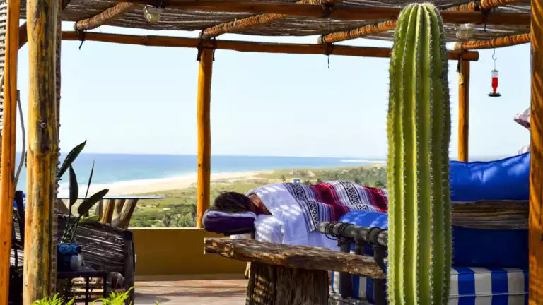 A guest relaxes on a massage table in the spa, with an outdoor patio, cacti, desert vegetation, and views of the Pacific Ocean, at Los Colibris Casitas, a boutique hotel in Baja.