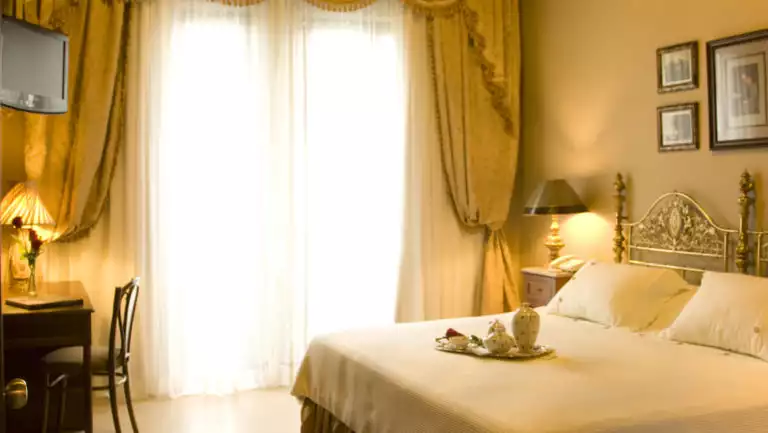 Soft light filters through curtains into a room with a queen-sized bed, white linens, and elegant furniture at Mansion del Rio, a premier boutique hotel in Guayaquil, Ecuador