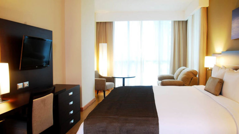 A room with a king-sized bed, television, chairs, and a large window at the Hotel Tryp by Wyndham Panama Centro