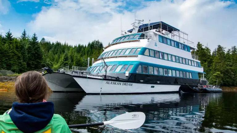 Kayaker paddling calm sea back to the 4-deck, blue & teal catamaran small ship Alaskan Dream on anchor by green forest.