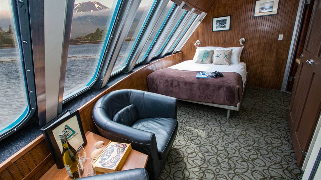 Vista View suite aboard Alaskan Dream with bed next to wall of windows and chair and table.