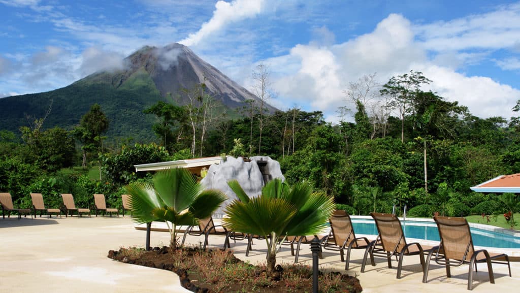 Costa Rica's Arenal Volcano rises behind a jungle forest, with the hotel's pristine pool, lounge chairs, and an umbrella in the foreground.