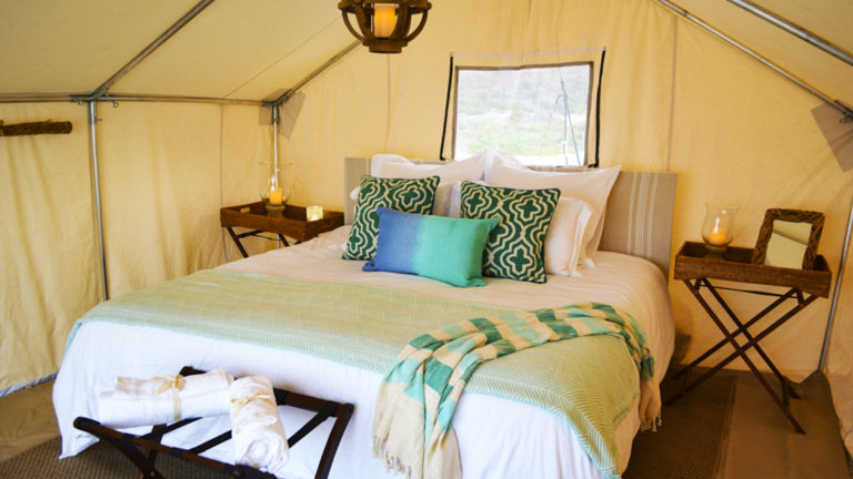 A queen-sized bed with blue accent pillows, bedside tables, and fresh linens in one of the glamping tents at Camp Cecil de la Isla, a beachfront abode on an island in Baja