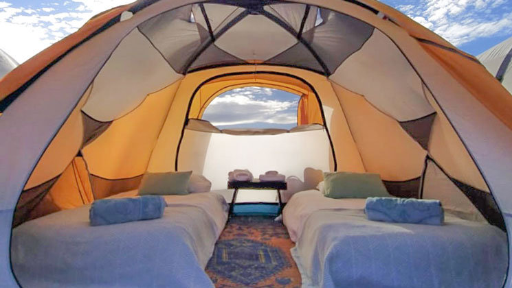 Interior of yellow & white dome tent with 2 air beds with sheets & blankets & bedside table for glamping Baja California.