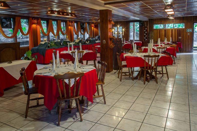 Round tables are set with red tablecloths, fine dining placements, and home-style wooden chairs at the restaurant inside Hotel Heliconia in Costa Rica's Monteverde region