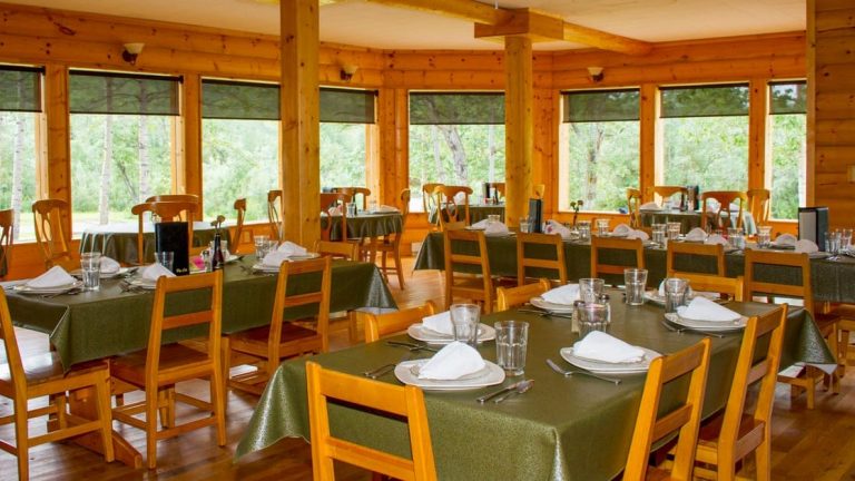 Dining area with wooden tables and chairs and large Bay windows at Kantishna Roadhouse Lodge in Alaska