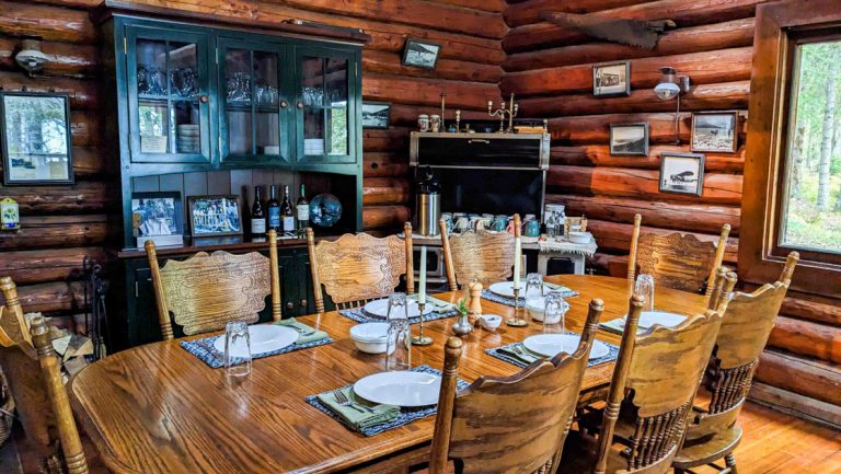 Dining room at Kenai Backcountry Lodge in Alaska with wood table set for 6, log cabin walls & dark green curio cabinet,