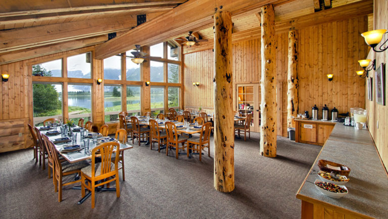 Long wooden tables and chairs are set for a meal inside a room with log cabin architecture and big windows looking toward the forest at the Kenai Fjords Glacier Lodge in Alaska