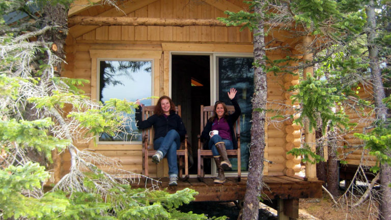 Two people sit on chairs on a covered patio just outside one of the private log cabins at the sustainable, eco resort Kenai Fjords Glacier Lodge in Alaska