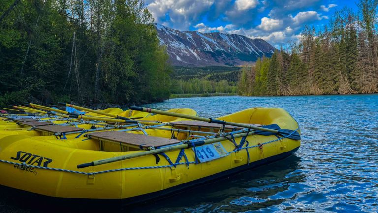 4 yellow rafts ready to go downriver sit on shore with green forest & tall mountains behind at Kenai Riverside Lodge.