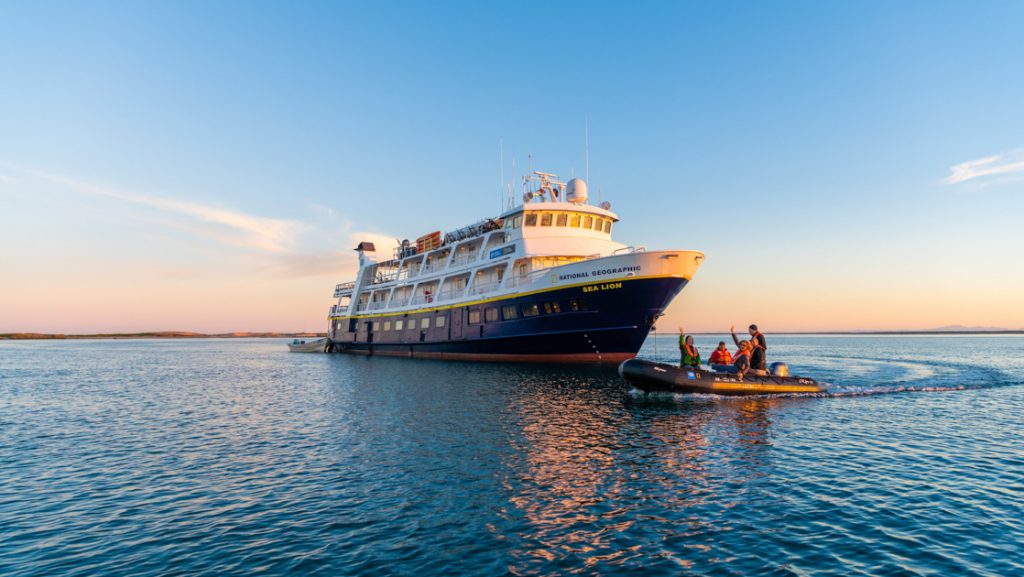National Geographic Sea Lion small ship with dark blue hull & white upper decks sits on anchor as Zodiac cruises by at dusk.