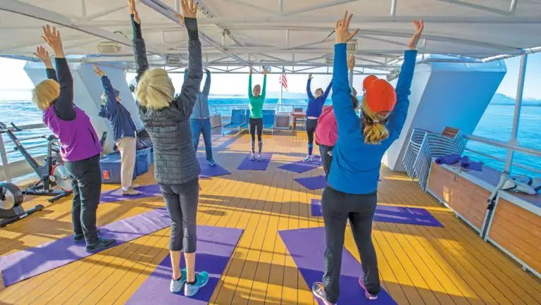 Guests stand & raise arms during morning stretch class atop purple yoga mats on deck of Nat Geo Sea Lion small ship.
