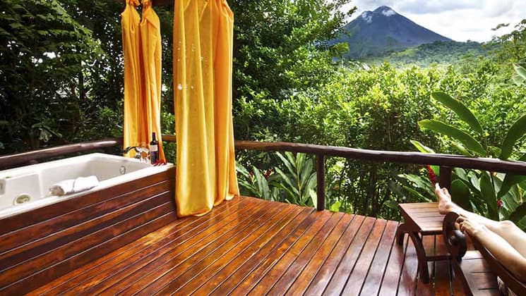 A person rests their legs on an ottoman on the deck, next to a jacuzzi, with a view of the Arenal Volcano National Park, adjacent to the Nayara Hotel, Spa & Gardens, a luxury boutique hotel in Costa Rica