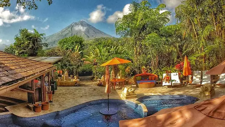 The pool, with umbrellas and lounge chairs, is set in a stone patio on the edge of the jungle with a view of the volcano at Nayara Hotel, Spa & Gardens, a luxury boutique hotel in Costa Rica