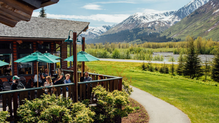Green umbrellas are opened on the deck outside of the Seward Windsong Lodge, while a path winds around the building through gardens and toward Alaska's snow-capped mountains
