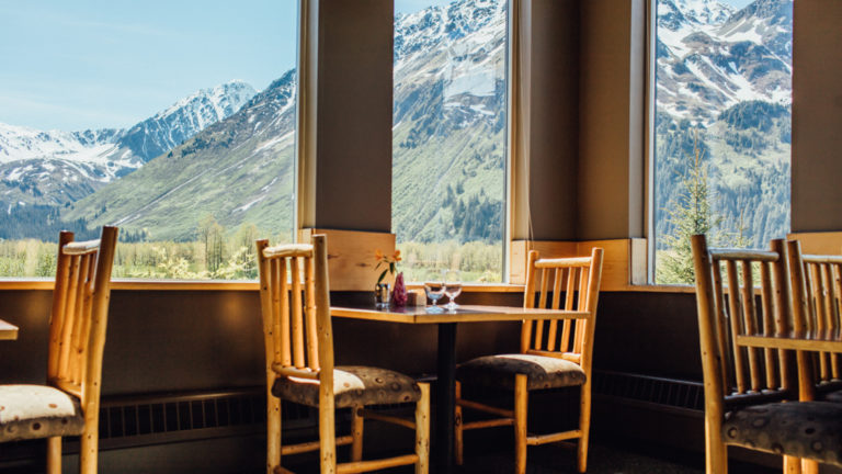 Tables and chairs next to large windows facing a view of the snow-capped mountains at Seward Windsong's gourmet restaurant, Resurrection Roadhouse, in Alaska
