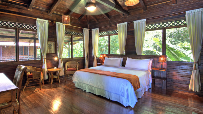 An upstairs balcony room with a king-sized bed, curtains, windows, tables, and chairs at the Tortuga Lodge, a luxury sustainable resort in Costa Rica.