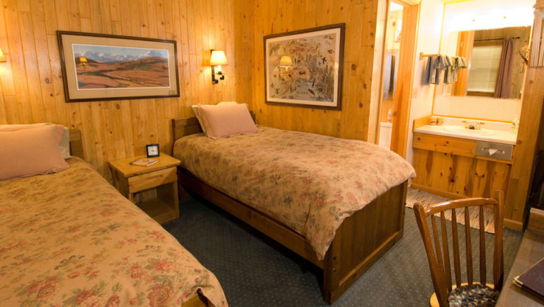 A room with two twin beds, artwork, and wood walls at the North Face Lodge, a traditional inn located in the Alaskan wilderness within Denali National Park
