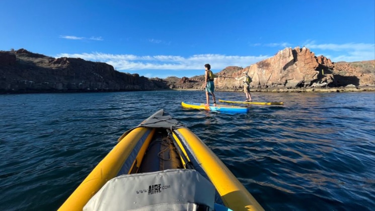 View from an inflatable yellow kayak, watching stand-up paddlers move in calm water towards sandy rock outcrops in Baja.