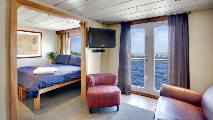 Safari Endeavour Alaska small ship Commodore suite with a large open room with glass doors leading to a balcony, a tv and a large bed in the adjoining room
