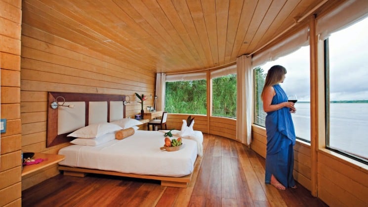 Woman standing in Master Suite with large bed and 180-degree picture windows aboard Delfin II riverboat on Amazon River cruise