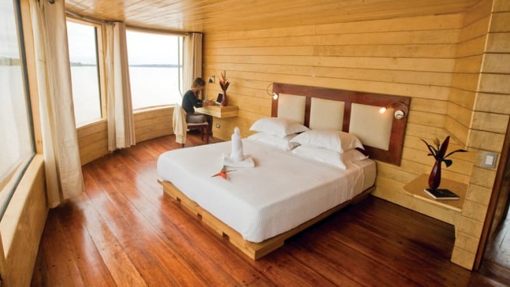 Woman writes at corner desk in Master Suite with large bed and picture windows aboard Delfin II riverboat on Amazon River cruise