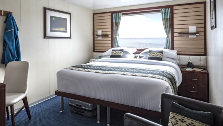 Category 4 cabin interior with large bed, armchair, bedside tables, desk, chair and window aboard National Geographic Endeavour II expedition ship in the Galapagos Islands