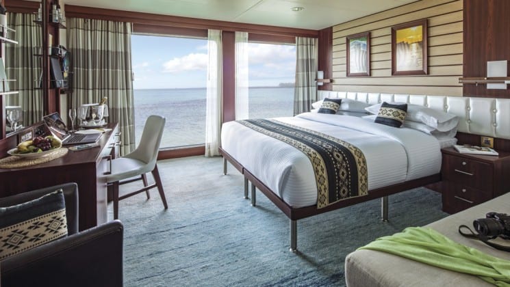 Interior of Suite B with large bed, couch, armchair, desk, chair and floor-to-ceiling windows aboard National Geographic Endeavour II expedition ship in the Galapagos Islands