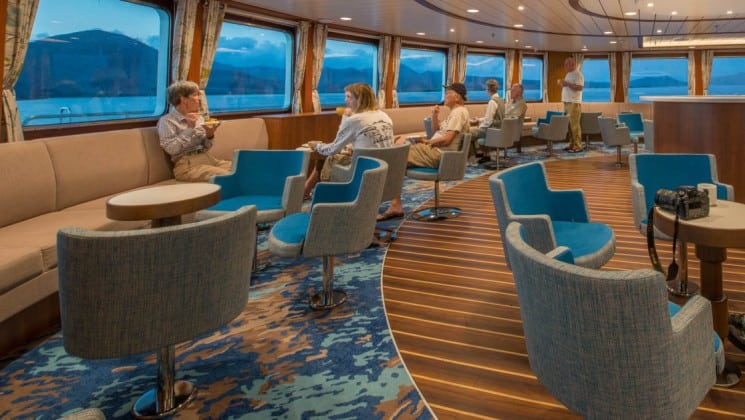 Passengers sit at couches and tables beneath large windows in the lounge aboard National Geographic Endeavour II expedition ship in the Galapagos Islands