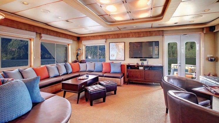 the lounge aboard Safari Quest Alaska small ship, with a coffee table and ottomans, windows, couch with pillows and a table and chairs