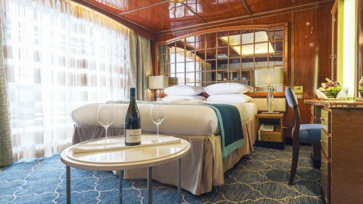 Wine bottle and glasses on small table beside bed, desk and chair in Deluxe Suite aboard Sea Spirit small ship