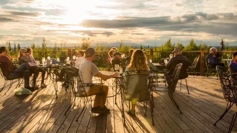 A large number of guests sitting on the deck watching the sun set at tables with food and drink at Talkeetna Lodge in Alaska.