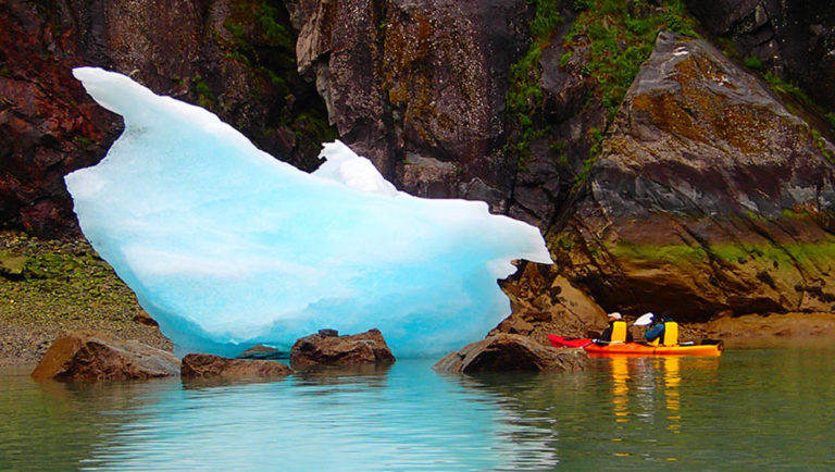 Tandem kayak couple up close to a blue iceberg floating close to rocks on the shore in Alaska.