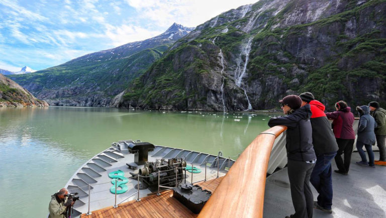 Passengers on a small ship enjoying the view from a deck above the bow as they are cruising past waterfalls coming down the fjords in Alaska.