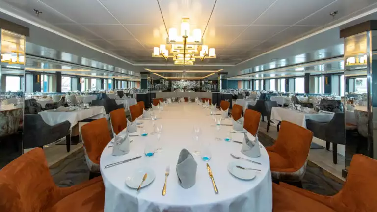 Long table set for formal dinner with white tablecloth, fine silverware, folded napkins and orange chairs aboard World Explorer polar small ship.