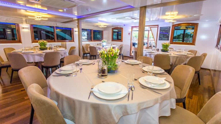 Dining Room aboard Aquamarin Croatia small ship, with small round tables set with white plates, beige tablecloth & tulips in a vase, wooden floor & buffet bar.