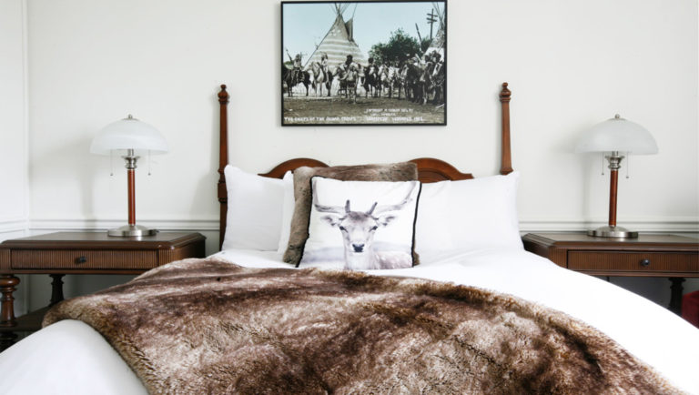Guest room double bed with white duvet, arctic reindeer-graphic throw pillow, animal skin throw blanket & 2 side tables at Fort Garry Hotel in Churchill, Canada.