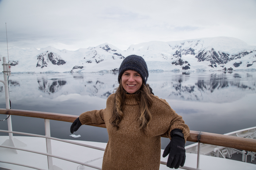 Woman in brown sweater, black gloves & blue knit hat stands on deck of small ship with snowy peaks behind in Antarctica.