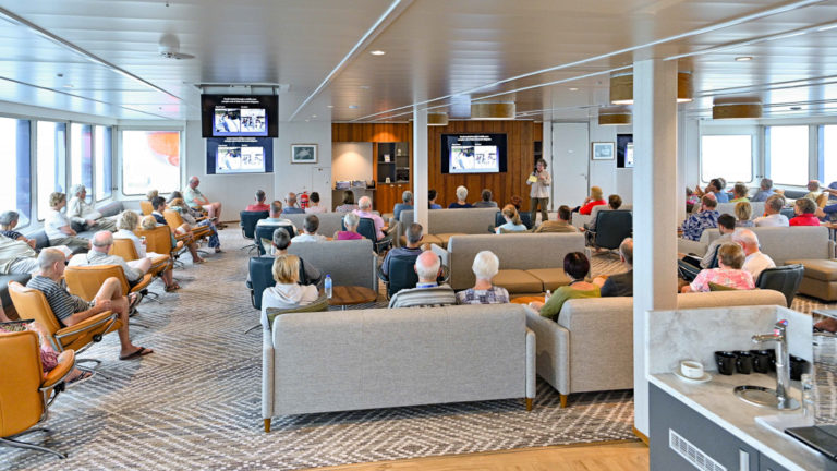 Lecture in the bridge deck lounge with sofas and tv screens aboard Coral Adventurer.