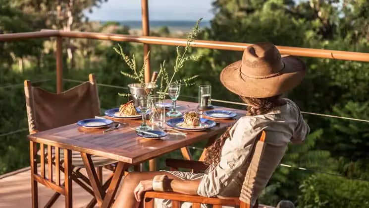Woman with wide-brim hat sitting at table on outside deck with forest and ocean views at Galapagos Safari Camp Santa Cruz Highlands in the Galapagos Islands