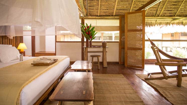 A bed with linens, wood furniture, open windows, and a mosquito net are inside the Amazonica Suite, a rustic comfort option at Inkaterra Reserva Amazonica