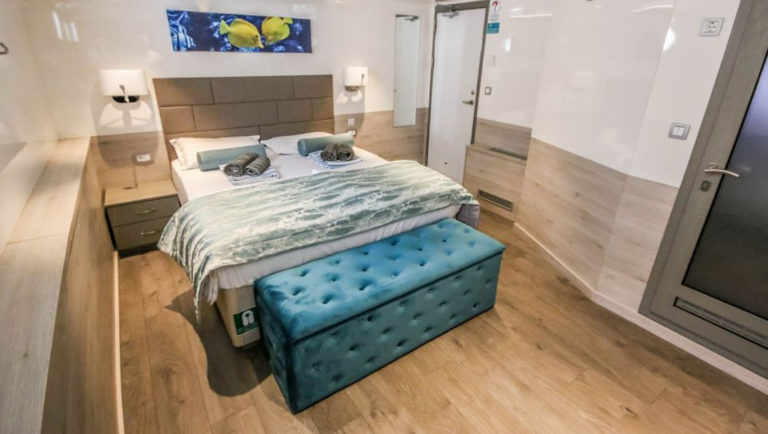 Guest cabin with double bed, plush bedding, blue velvet storage chest, wooden accents, shiny white walls and two bedside tables with lamps aboard Nautilus Croatia & Mediterranean deluxe small yacht.