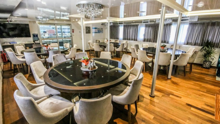 Dining Room with black-and-gold circular tables surrounded by gray velour chairs, wooden floors, crystal chandeliers, a TV on the wall, silver support poles & bright white lighted ceiling aboard Nautilus Croatia & Mediterranean deluxe small yacht.